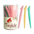 A clear cylindrical container of Tinkle Eyebrow Rom with a cream-colored label featuring a red-haired cartoon woman and the brand name 'Tinkle'. Three eyebrow grooming tools are displayed outside the container: one pink, one pale yellow, and one mint green, all on a clean white background.