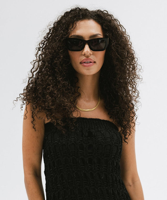 Woman wearing black sunglasses, a gold chain and a black top against a plain background  #color_black