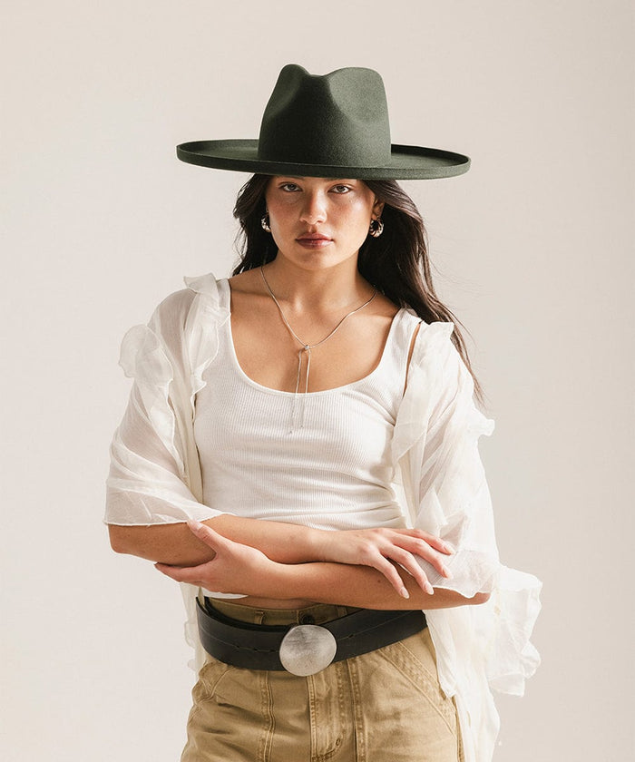 Woman wearing a dark green pencil brim fedora hat and a white top with a  white blouse against a plain background #color_dark green