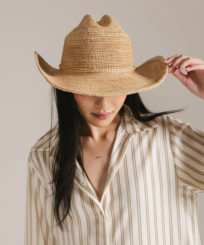 Woman wearing a natural colored straw cowboy hat and a light colored shirt, looking down against a plain background #color_natural