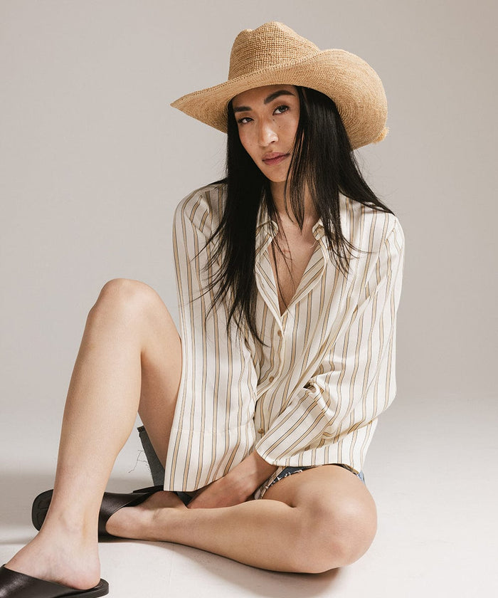 Woman wearing a natural colored straw cowboy hat, a light colored shirt and brown slippers, sitting against a plain background #color_natural