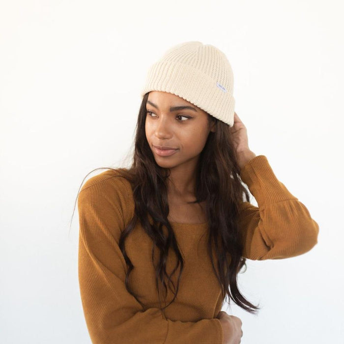 Woman wearing an oatmeal colored knit beanie with the brand label and a brown sweatshirt, looking sideways against a plain background #color_oatmeal