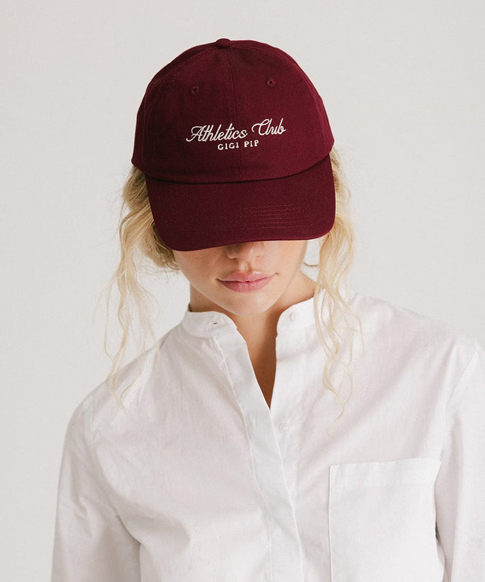 Woman wearing a burgundy cap with white 'Athletics Club' text and a white shirt against a plain background  #color_burgundy