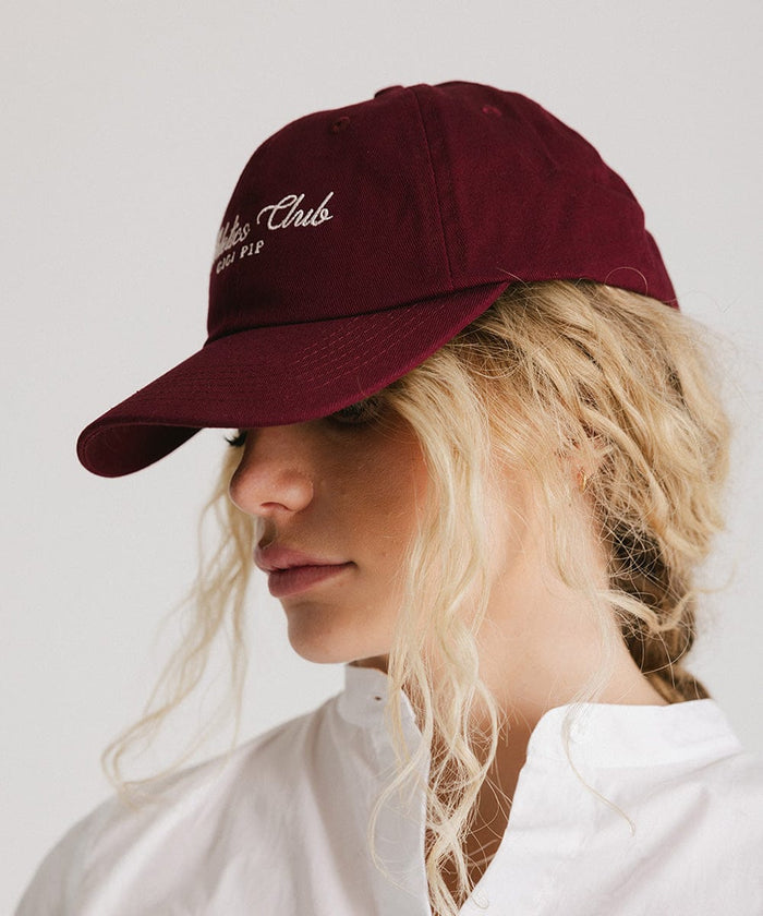Woman wearing a burgundy cap with white 'Athletics Club' text and a white shirt, posing sideways against a plain background  #color_burgundy