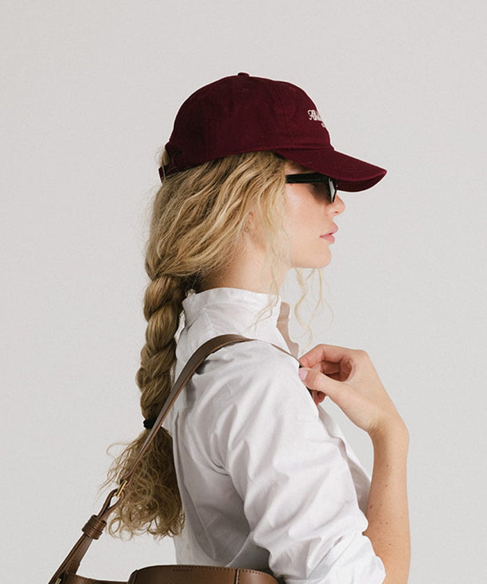 Woman wearing a burgundy cap, sunglasses and a white shirt, facing sideways against a plain background  #color_burgundy