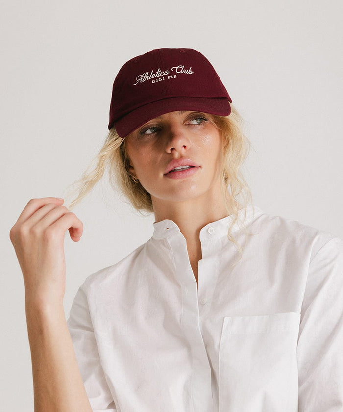 Woman wearing a burgundy cap with white 'Athletics Club' text and a white shirt against a plain background  #color_burgundy