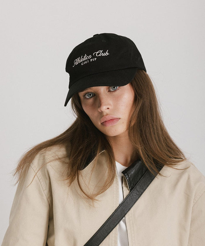 Woman wearing a black cap with white 'Athletics Club' text and a beige jacket against a plain background  #color_black