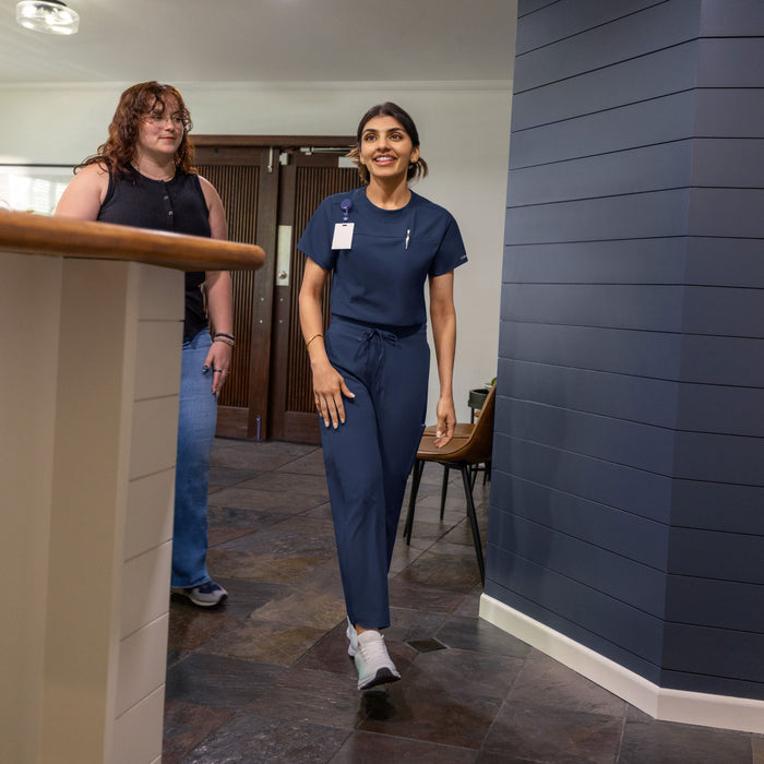 A woman in navy blue medical scrubs walks down a hallway in a hospital or clinic setting, while another person stands behind a counter in the background.