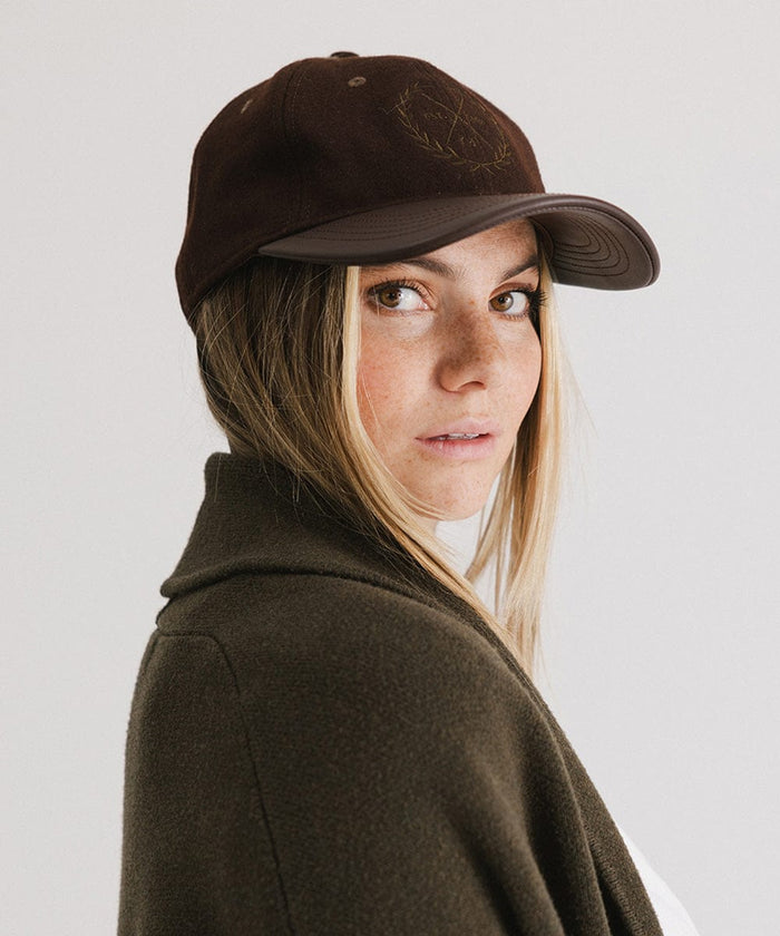 Woman wearing a brown cap with a logo and a white t-shirt with a brown coat, posing sideways against a plain background  #color_brown
