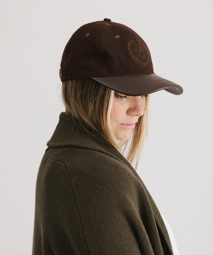Woman wearing a brown cap with a logo and a white t-shirt with a brown coat, facing sideways against a plain background  #color_brown