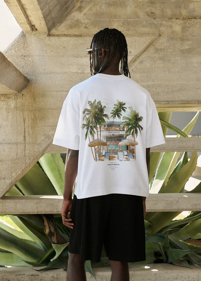 Person wearing a white t-shirt with a tropical design, standing in front of a concrete structure with plants.