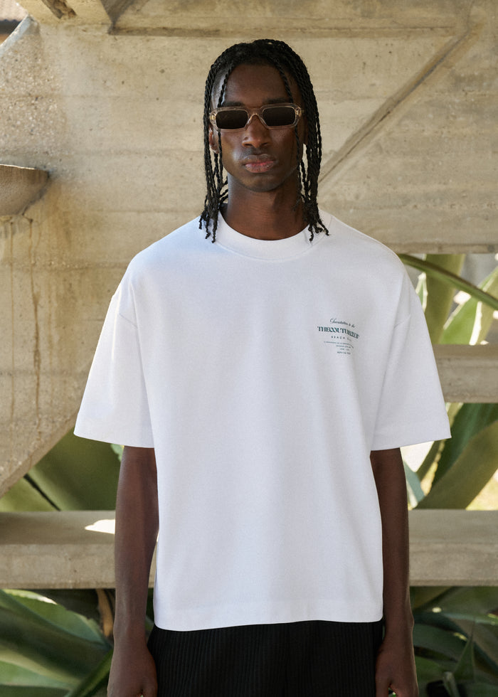 Man wearing a white t-shirt with a logo, standing outdoors with greenery and concrete structure in the background.