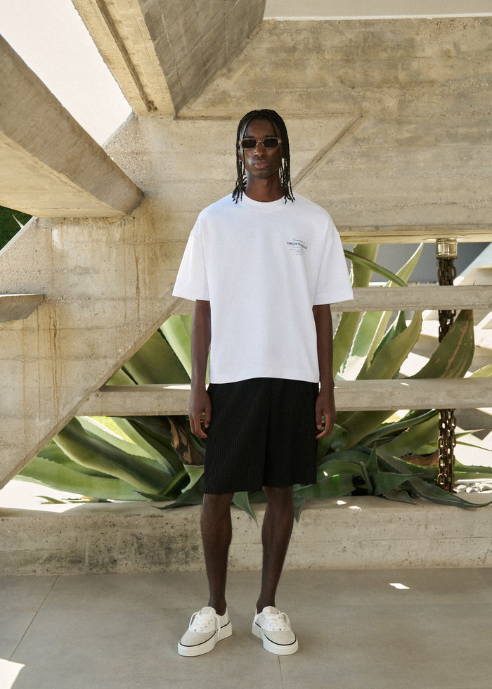 Person wearing a white t-shirt and black shorts standing in front of a concrete structure with plants.