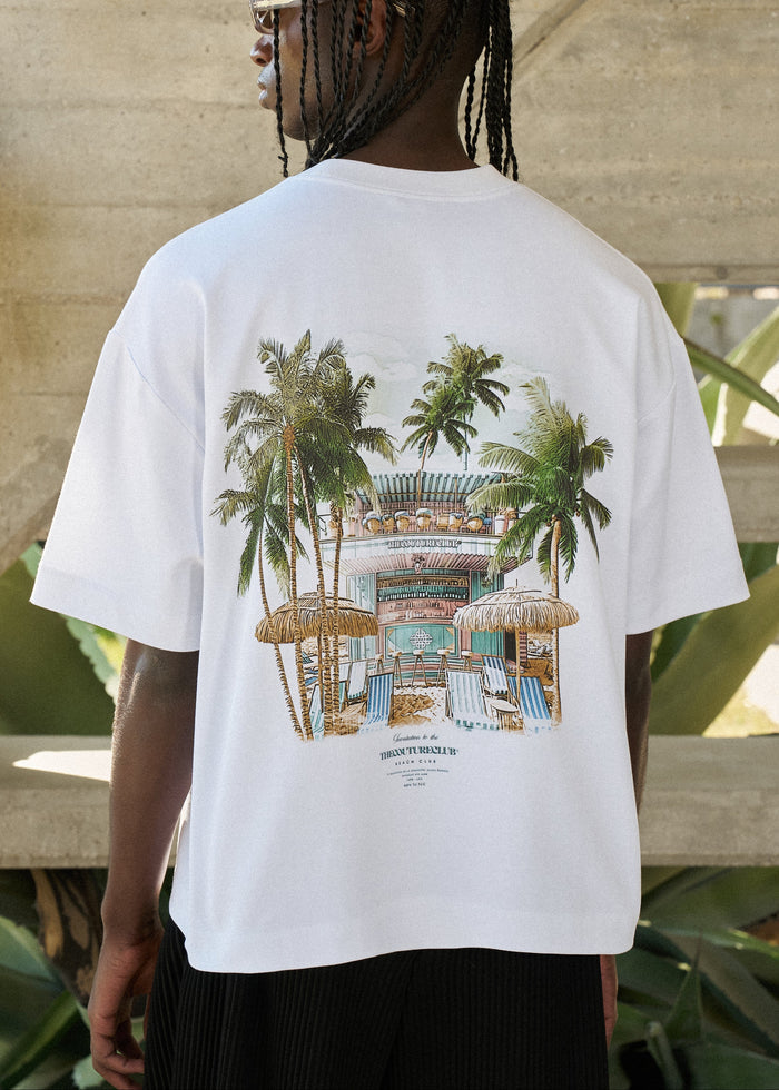 Person wearing a white t-shirt with a tropical beach club graphic on the back, standing outdoors.