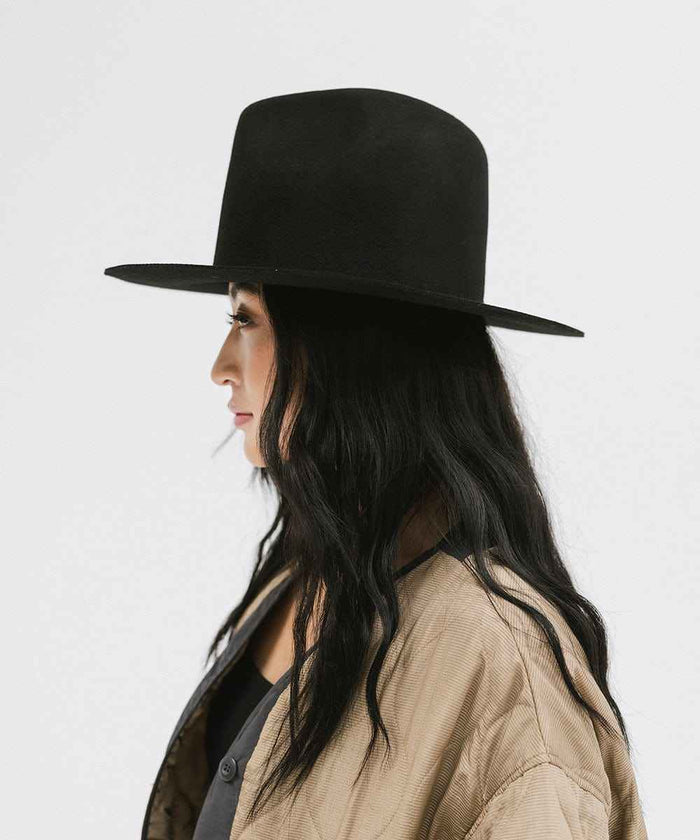 Woman wearing a black tall fedora hat and a beige jacket, facing sideways against a plain background #color_black