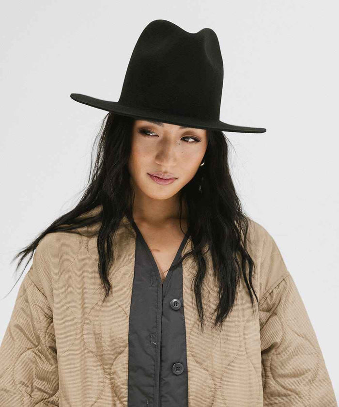 Woman wearing a black tall fedora hat and a beige jacket, smiling against a plain background #color_black