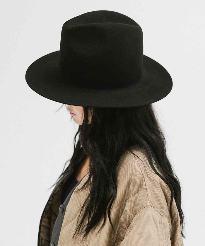 Woman wearing a black tall fedora hat and a beige jacket, posing sideways against a plain background #color_black