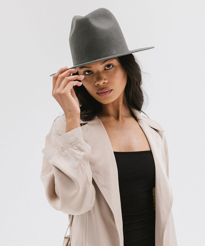 Woman wearing a dark grey tall fedora hat and a black top with a beige coat against a plain background #color_dark grey