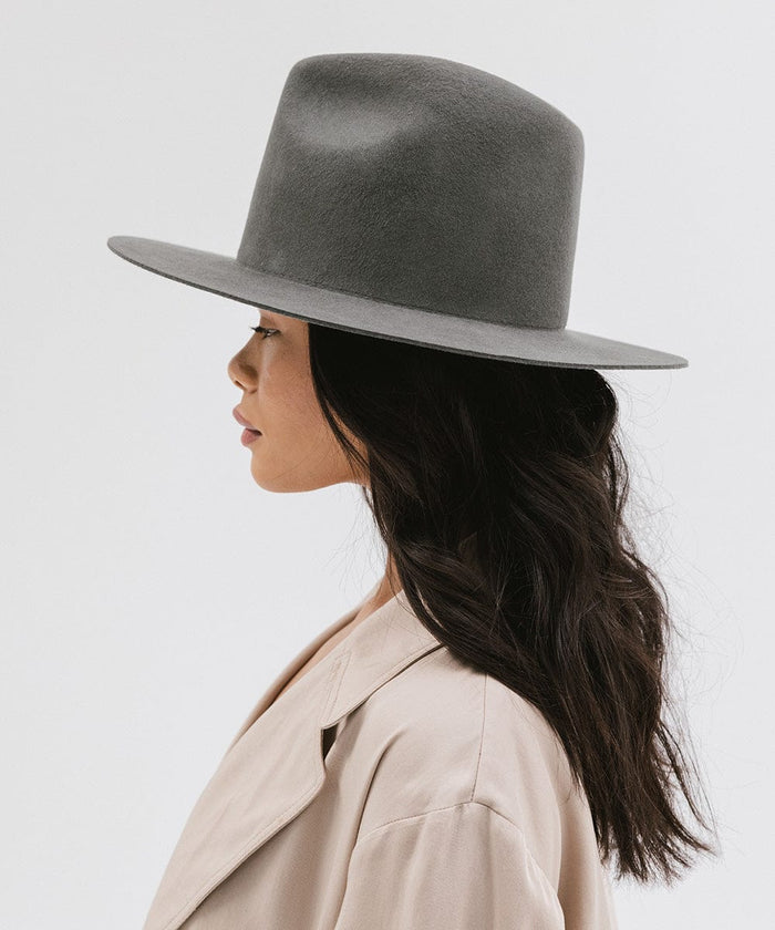 Woman wearing a dark grey tall fedora hat and a beige coat, facing sideways against a plain background #color_dark grey