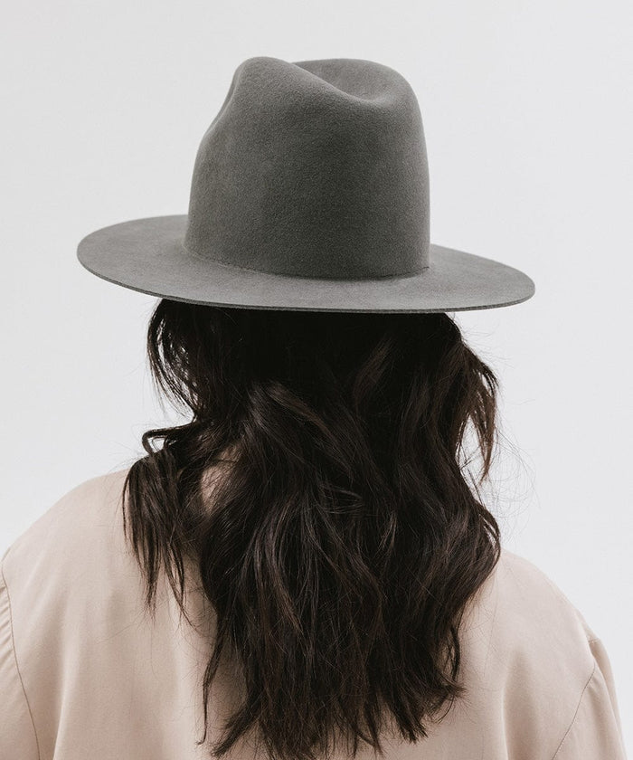 Woman wearing a dark grey tall fedora hat and a beige coat, facing behind against a plain background #color_dark grey
