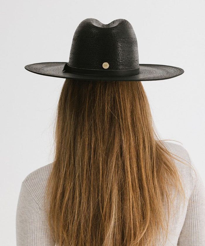 Woman with long hair wearing a black wide brim fedora hat with a hat band facing behind against a plain background #color_limited edition black