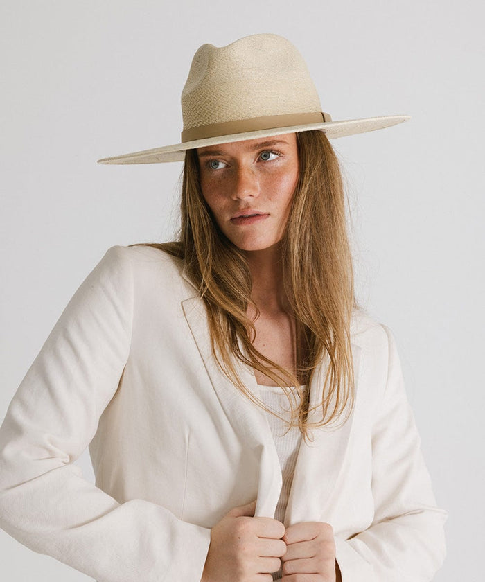 Woman wearing a natural colored wide brim fedora hat with a hat band and a cream blazer against a plain background #color_natural