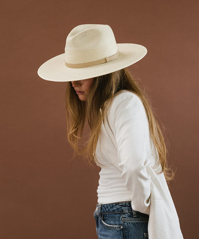 Woman wearing a natural colored wide brim fedora hat with a hat band and a cream blazer, posing against a brown background #color_natural