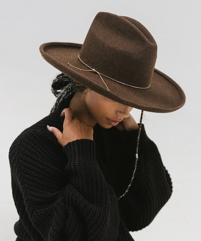 Woman wearing a brown hat with a gold chain bolo hat band and a black sweater, posing sideways against a plain background  #color_gold