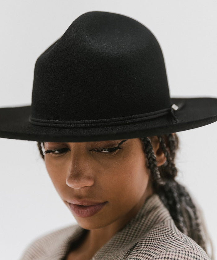 Close up image of a woman wearing wearing a black fedora hat with a hat band against a plain background #color_black