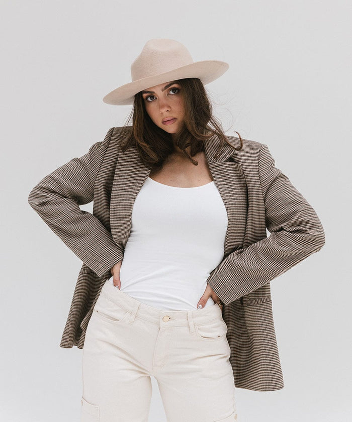 Woman wearing a cream fedora hat with a hat band, a white top with a checkered coat and white pants against a plain background #color_cream