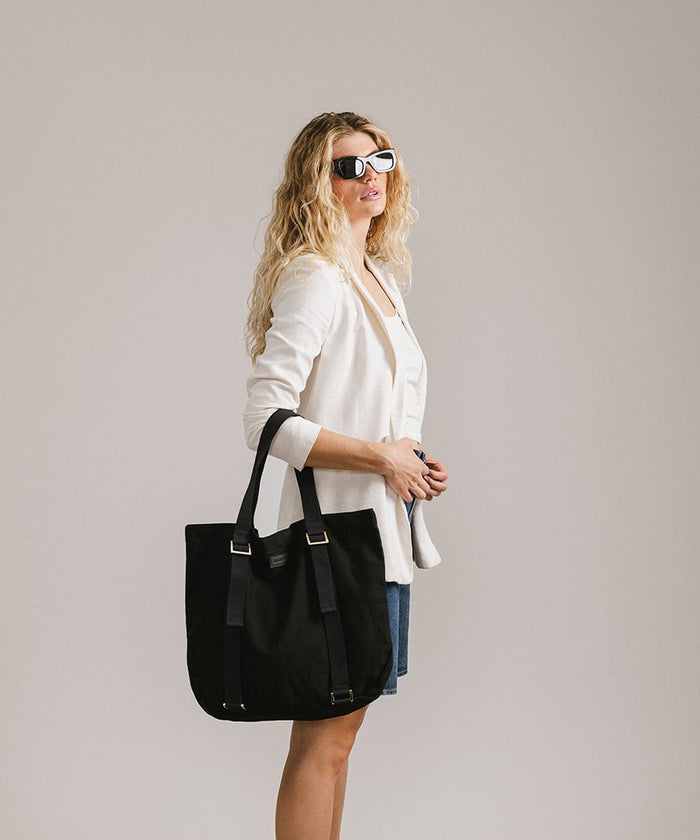 Woman wearing sunglasses, a white top with a white jacket and denim shorts, carrying a black canvas tote bag facing sideways against a plain background #color_black