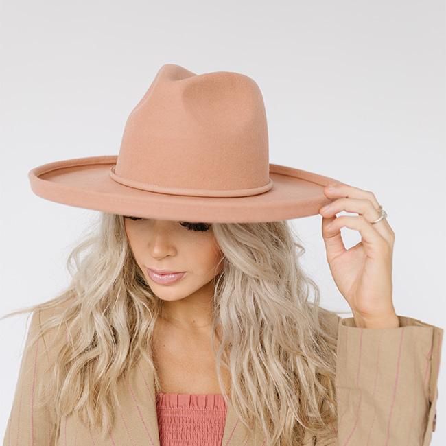 Woman wearing a dusty pink pencil brim fedora hat and a beige blazer, looking down against a plain background  #color_dusty pink