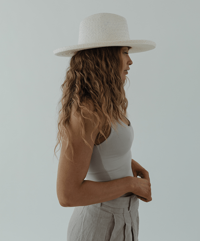 Woman wearing a white pencil brim straw hat, a white top and grey pants, facing sideways against a plain background #color_white