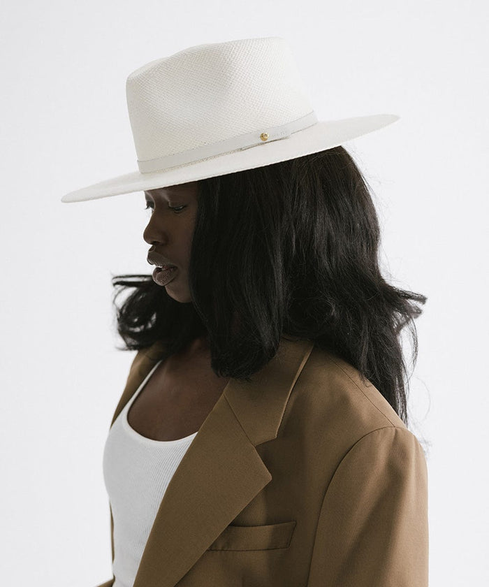 Woman wearing a white straw hat with a hat band and white top with a brown coat, facing sideways against a plain background #color_white