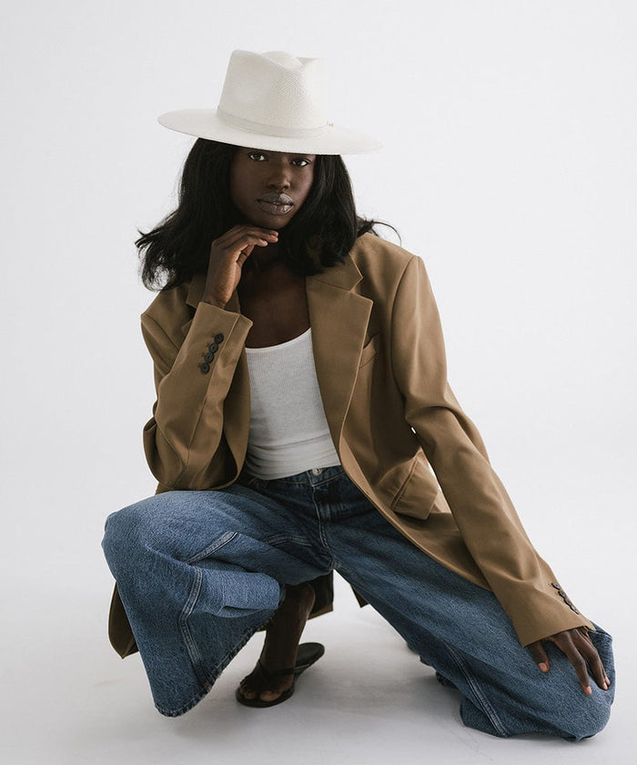 Woman wearing a white straw hat with a hat band, a white top with a brown coat, and blue jeans, sitting against a plain background #color_white