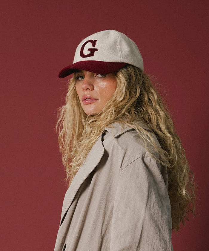 Woman wearing a white and burgundy baseball cap with the letter 'G' and a beige shirt against a red background  #color_burgundy