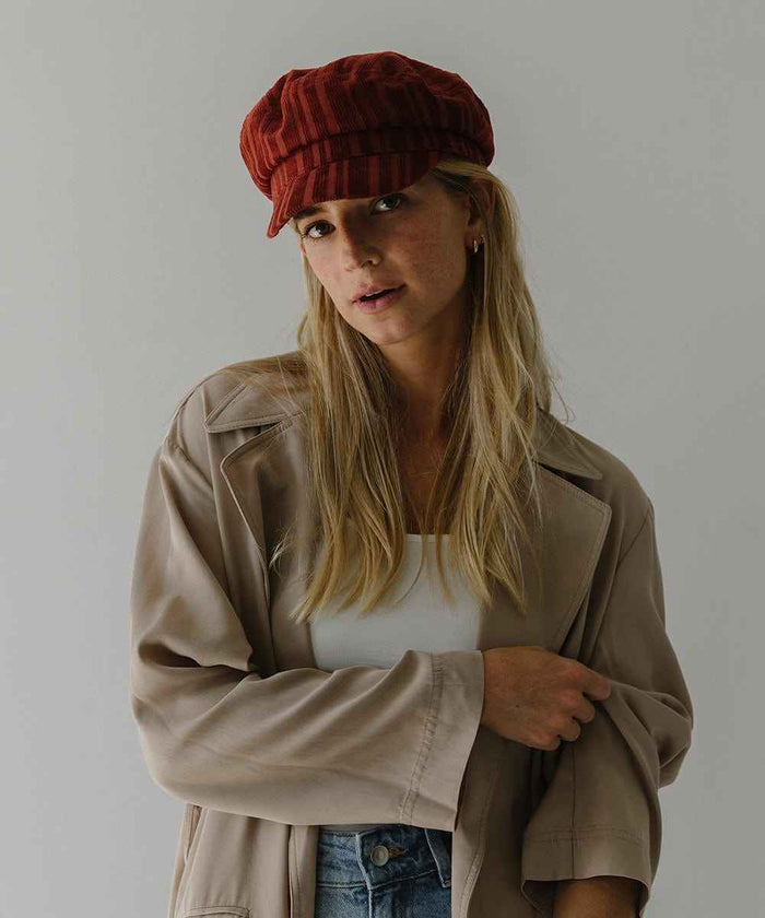 Woman wearing a crimson red colored striped cap and a white top with a beige jacket against a plain background #color_crimson red
