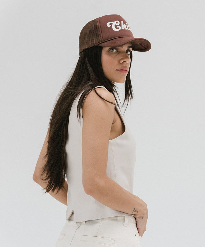 Woman wearing a chocolate brown colored trucker hat with the text 'Chill' and a white top, posing against a plain background #color_chocolate brown