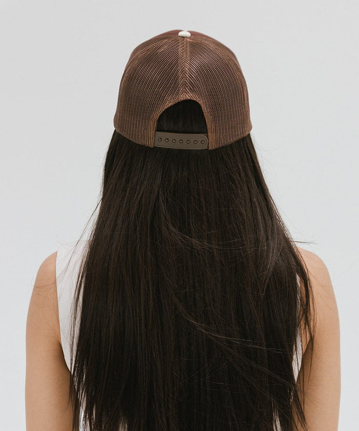 Woman wearing a chocolate brown colored trucker hat and a white top, facing behind against a plain background #color_chocolate brown