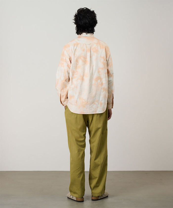 Back view of a model wearing a peach Bleach Stripe colored Gramicci Classic Oxford Bleached Shirt and green pants on a light-tone background.
