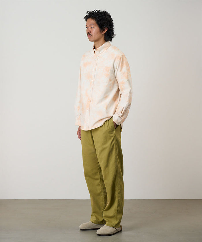 Model wearing a peach Bleach Stripe colored Gramicci Classic Oxford Bleached Shirt and green pants on a light-tone background.