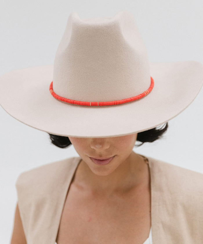 Woman wearing a cream hat with a red beaded hat band and a beige top against a plain background #color_red