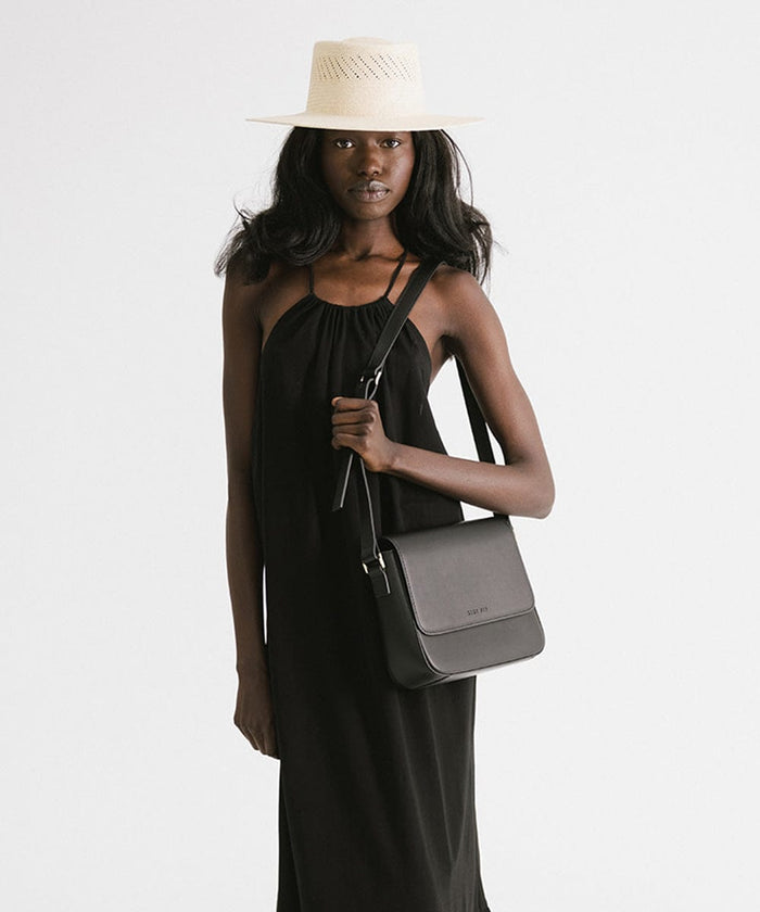 Woman wearing a natural colored straw hat and a black top, carrying a black bag against a plain background #color_natural