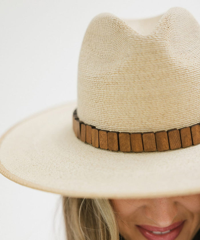 Person wearing a natural colored wide brim straw hat with a brown hat band against a plain background  #color_natural