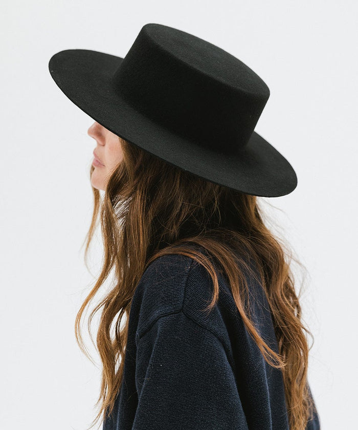 Woman with long hair wearing a black boater-style hat and a dark blue shirt, facing sideways against a plain background #color_black