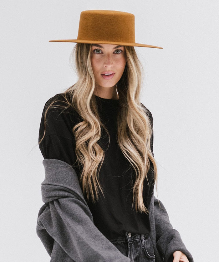 Woman with long hair wearing a brown boater-style hat, a black t-shirt and grey jacket against a plain background #color_brown