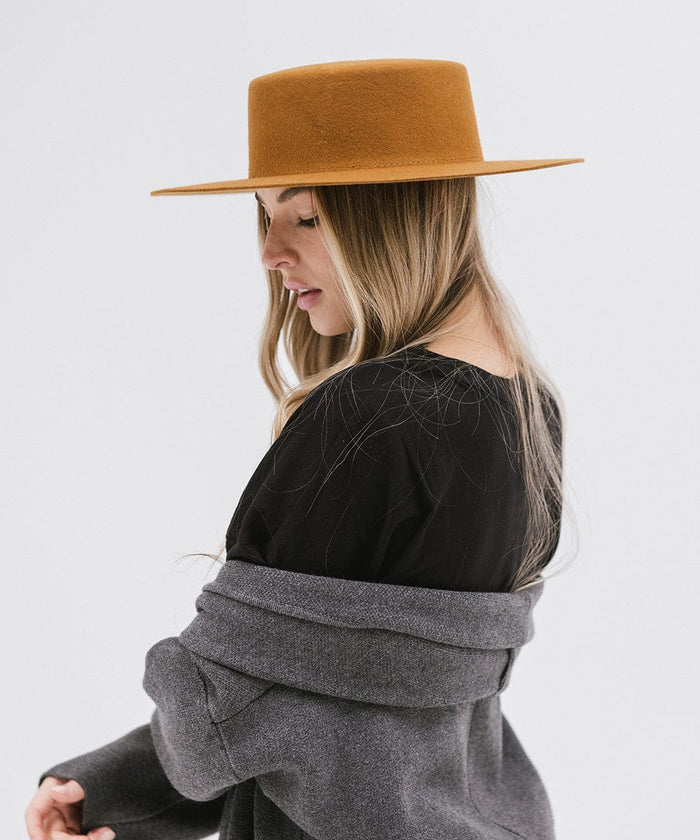 Woman wearing a brown boater-style hat, a black t-shirt and grey jacket, posing sideways against a plain background #color_brown