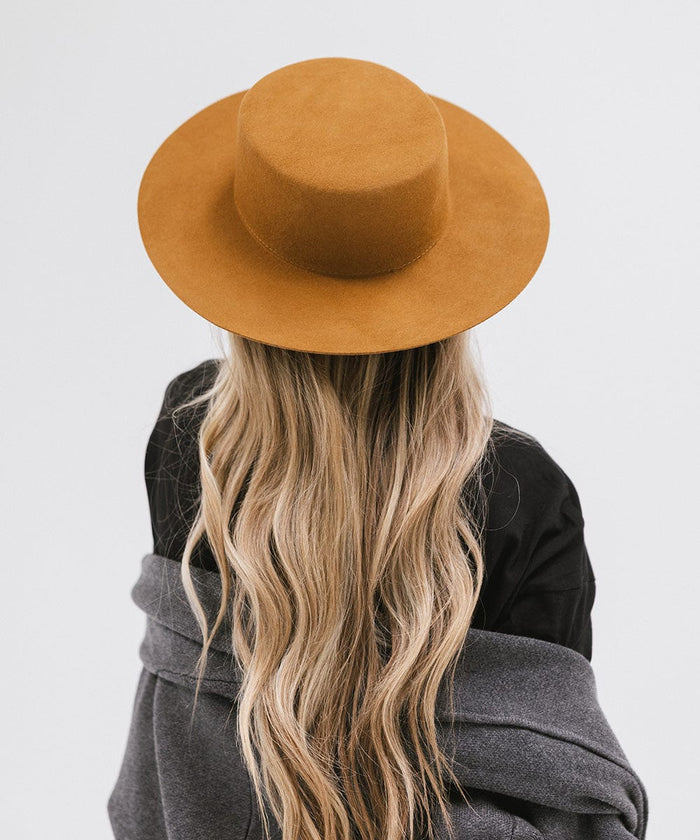 Woman long blonde hair wearing a brown boater-style hat, a black t-shirt and grey jacket, facing behind against a plain background #color_brown