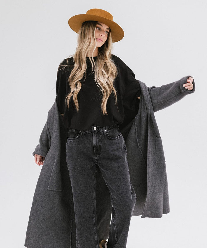Woman with long hair wearing a brown boater-style hat, a black t-shirt, grey jacket and dark grey jeans against a plain background #color_brown