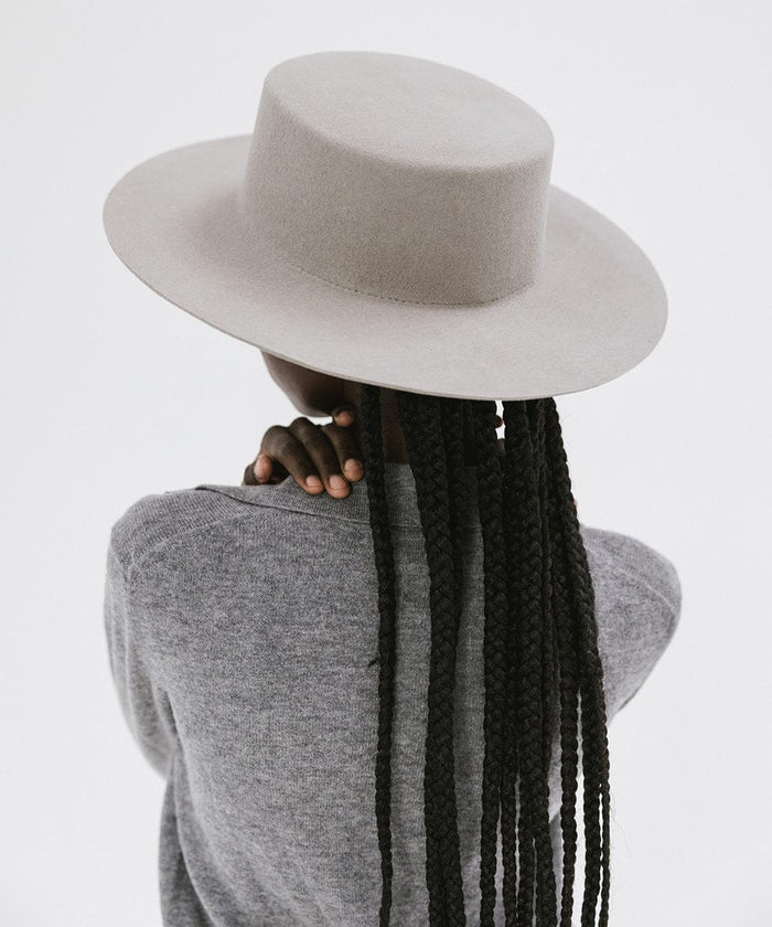 Woman with long hair wearing a light grey boater-style hat and a grey shirt, facing behind against a plain background #color_light grey